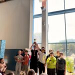Zdjęcia z ceremonii zakończenia zawodów pływackich dzieci i młodzieży. Uczestnicy odbierają medale i puchary oraz pozują na podium. W wydarzeniu biorą udział zawodnicy, organizatorzy i kibice zgromadzeni na pływalni.