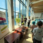 Zdjęcia z ceremonii zakończenia zawodów pływackich dzieci i młodzieży. Uczestnicy odbierają medale i puchary oraz pozują na podium. W wydarzeniu biorą udział zawodnicy, organizatorzy i kibice zgromadzeni na pływalni.