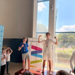 Zdjęcia z ceremonii zakończenia zawodów pływackich dzieci i młodzieży. Uczestnicy odbierają medale i puchary oraz pozują na podium. W wydarzeniu biorą udział zawodnicy, organizatorzy i kibice zgromadzeni na pływalni.