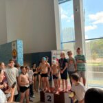 Zdjęcia z ceremonii zakończenia zawodów pływackich dzieci i młodzieży. Uczestnicy odbierają medale i puchary oraz pozują na podium. W wydarzeniu biorą udział zawodnicy, organizatorzy i kibice zgromadzeni na pływalni.