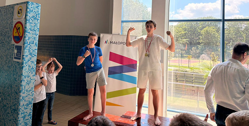 Zdjęcia z ceremonii zakończenia zawodów pływackich dzieci i młodzieży. Uczestnicy odbierają medale i puchary oraz pozują na podium. W wydarzeniu biorą udział zawodnicy, organizatorzy i kibice zgromadzeni na pływalni.
